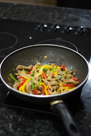 A frying pan on a stovetop contains a stir-fried dish with sliced beef, red and yellow bell peppers, and green vegetables. It is positioned on a dark countertop with a blurred background.