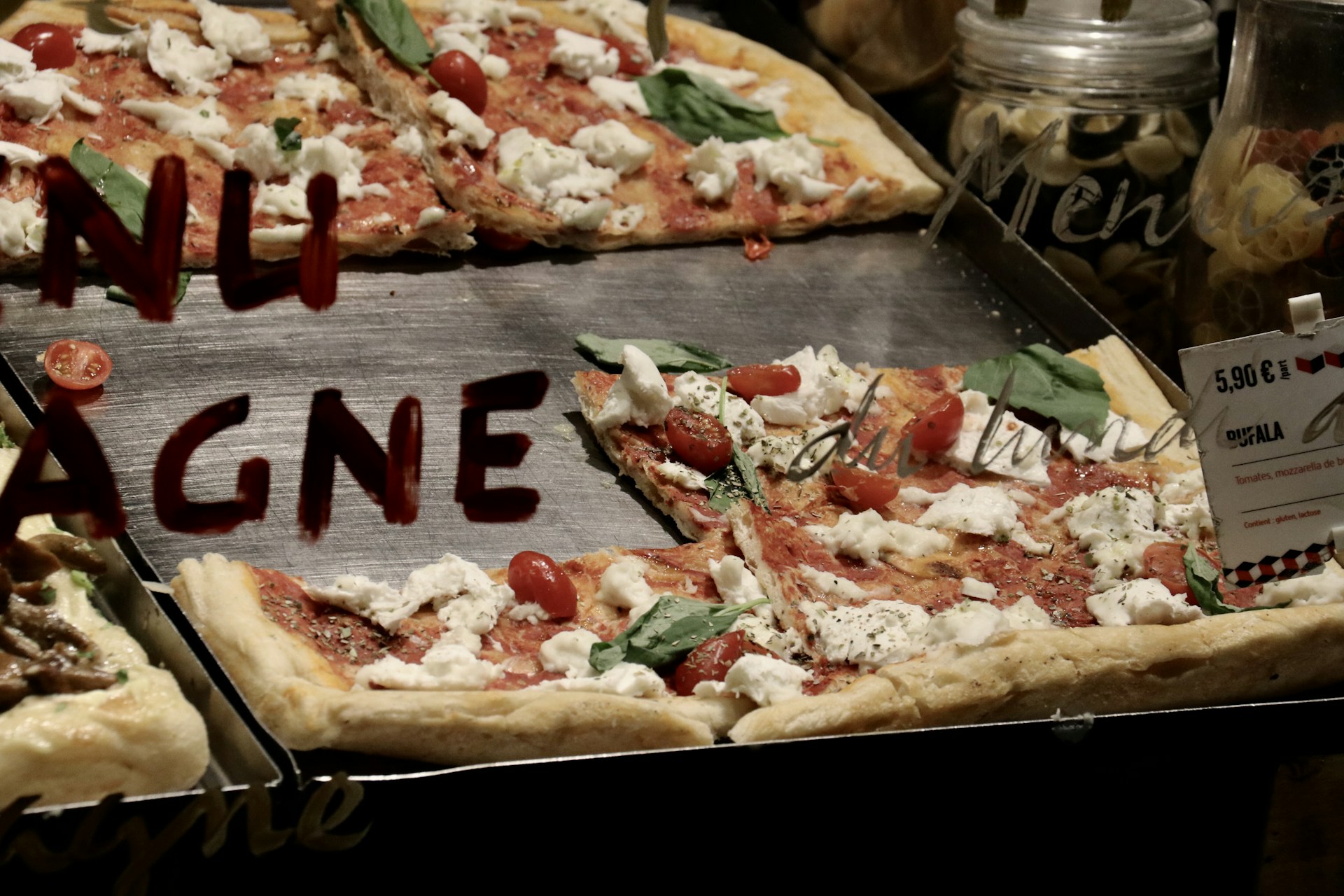 Slices of pizza topped with mozzarella cheese, cherry tomatoes, and basil leaves are displayed on a metal tray. The word 'TENTAZIONE' is partially visible on a glass surface positioned in front of the pizza. There's also a price tag placed on the tray, indicating the cost of 5.90 Euros.