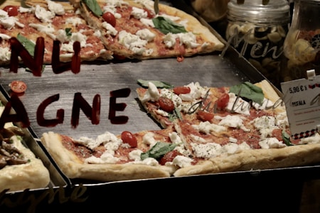 Slices of pizza topped with mozzarella cheese, cherry tomatoes, and basil leaves are displayed on a metal tray. The word 'TENTAZIONE' is partially visible on a glass surface positioned in front of the pizza. There's also a price tag placed on the tray, indicating the cost of 5.90 Euros.