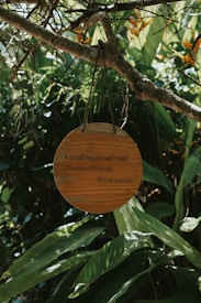 A round wooden sign hangs from a tree branch in a lush, green environment filled with large leaves and plants. The sign bears the inscription, 'A small house will hold a hundred friends. - African proverb'. The natural surroundings give a feeling of tranquility and connectedness with nature.