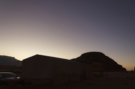 A desert landscape at dusk features a large tent with a sloped roof and a vehicle parked nearby. Silhouetted against the fading light, a large rock formation looms in the background. The sky transitions from a pale yellow at the horizon to a deep blue at the top, with a faint crescent moon visible.