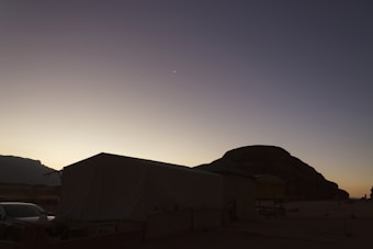 A desert landscape at dusk features a large tent with a sloped roof and a vehicle parked nearby. Silhouetted against the fading light, a large rock formation looms in the background. The sky transitions from a pale yellow at the horizon to a deep blue at the top, with a faint crescent moon visible.