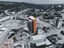 An aerial view of an urban landscape featuring a tall, modern building with orange accents. Surrounding the building are roads, a crane, and other smaller structures. The scenery includes more buildings in the background, hills, and greenery, under a cloudy sky.