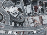 An aerial view captures an urban area featuring a complex of modern buildings, a construction site, and several roads. The scene includes multiple cars parked and in motion, as well as a crane at the construction site. The layout of the roads includes a roundabout and various intersections.