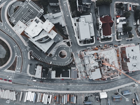 An aerial view captures an urban area featuring a complex of modern buildings, a construction site, and several roads. The scene includes multiple cars parked and in motion, as well as a crane at the construction site. The layout of the roads includes a roundabout and various intersections.