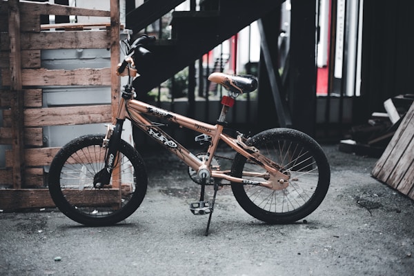A small bicycle with a tan and black color scheme is parked against a wooden pallet in an industrial or warehouse-like setting. The frame has various stickers, and it stands on a kickstand. The background includes dark stairs and wooden planks, contributing to an urban and gritty atmosphere.