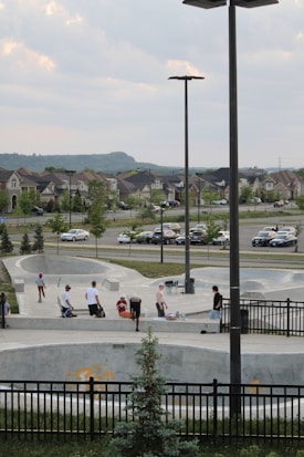 A skatepark scene with several people engaging in activities such as skateboarding and socializing. The foreground features a skateboard bowl with a black metal fence surrounding it. Behind the bowl, young people are standing and sitting near the edge, while a few are on their skateboards. In the background, there is a suburban neighborhood with houses, trees, and parked cars. The setting appears peaceful with cloudy skies overhead.