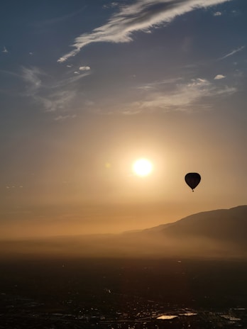 A hot air balloon floats gracefully against the backdrop of a serene sky during either sunrise or sunset. The sun casts a warm golden hue across the horizon, partially obscured by wispy clouds. Below, a landscape is faintly visible, shrouded in a gentle mist that enhances the tranquil atmosphere.
