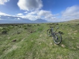 A mountain bike stands on a grassy trail that extends towards a distant range of mountains under a partly cloudy sky. The scene captures a sense of openness and adventure, with lush greenery and rugged terrain.