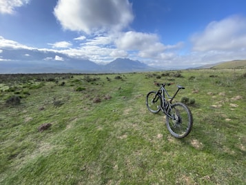 A mountain bike stands on a grassy trail that extends towards a distant range of mountains under a partly cloudy sky. The scene captures a sense of openness and adventure, with lush greenery and rugged terrain.