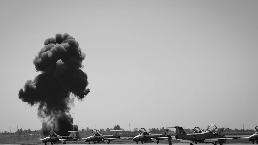 عمود دخان أسود كثيف يتصاعد خلف صف من الطائرات العسكرية على مدرج، بالأبيض والأسود