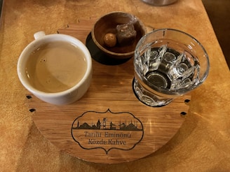 A wooden circular tray holds a cup of coffee, a small glass of water, and a bowl containing a piece of Turkish delight. The tray features a decorative design with text and skyline imagery.