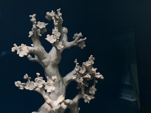 A white ceramic sculpture resembling a tree with delicate flowers on its branches. The background is a deep, dark blue, which contrasts with the light color of the sculpture. The tree-like structure and its intricate details create an elegant and peaceful composition.