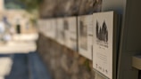 A series of information plaques mounted on a stone wall. The foreground is focused on one particular plaque displaying an image of trees and text in a language that appears to be Hebrew. The background is blurred, showing a continuation of the wall and some architectural elements in an outdoor setting.