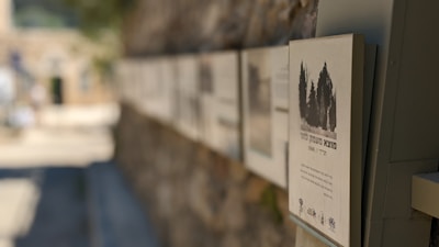 A series of information plaques mounted on a stone wall. The foreground is focused on one particular plaque displaying an image of trees and text in a language that appears to be Hebrew. The background is blurred, showing a continuation of the wall and some architectural elements in an outdoor setting.