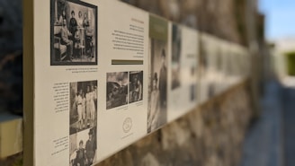 A long display of historical photographs and text is mounted along a textured stone wall. The images are likely black and white, some featuring group portraits and scenes from the past with accompanying text in a language possibly Hebrew. The setting is outdoors, with a blurred background suggesting depth and focus on the pictures.