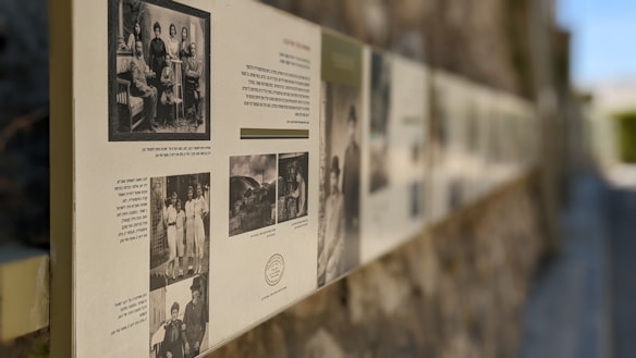 A long display of historical photographs and text is mounted along a textured stone wall. The images are likely black and white, some featuring group portraits and scenes from the past with accompanying text in a language possibly Hebrew. The setting is outdoors, with a blurred background suggesting depth and focus on the pictures.