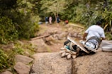 A pile of sandals and camping gear is placed on a rock in the foreground. In the background, a path leads into a dense forest with several people walking. The scene is natural and earthy, suggesting a hiking trip.