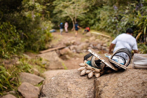 A pile of sandals and camping gear is placed on a rock in the foreground. In the background, a path leads into a dense forest with several people walking. The scene is natural and earthy, suggesting a hiking trip.