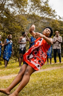 A person wearing a vibrant red traditional garment with black and white patterns appears to be dancing or performing a cultural gesture in a grassy outdoor setting. Surrounding this individual are several other people, dressed in blue and white traditional attire, standing in a circle. The background consists of green trees and hills.