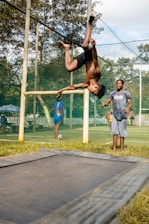 A person is performing a backflip on a trampoline in an outdoor setting. Two other individuals are observing nearby, and a soccer ball is visible on a grass field surrounded by a net. Trees and a clear sky form the background.