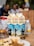 Three cartons of beverage labeled 'Orange Grove Amahewu asemZansi cool cream' are placed on a table. The cartons feature silhouettes of people dancing and running. In the foreground, there is a plate of food, including rice and a visible cucumber slice. An intricately designed glass is nearby, along with a plastic bottle in the background. Blurred figures and objects are visible in the background, suggesting a busy setting.