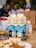 Three cartons of beverage labeled 'Orange Grove Amahewu asemZansi cool cream' are placed on a table. The cartons feature silhouettes of people dancing and running. In the foreground, there is a plate of food, including rice and a visible cucumber slice. An intricately designed glass is nearby, along with a plastic bottle in the background. Blurred figures and objects are visible in the background, suggesting a busy setting.