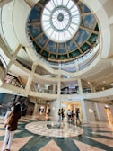 A large, modern shopping mall interior with multiple floors visible. The ceiling is an ornate, circular glass dome with a blue sky and clouds painted on it. Shoppers are walking around the open space, and there are stores like Dior and Guess visible. The floor has a decorative circular pattern.