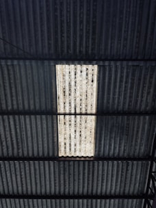 A corrugated metal roof with a rectangular section that is translucent or a different color, allowing light to pass through. The roof structure includes multiple lines and patterns in its design, with some areas appearing worn or weathered.
