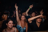 A crowded club scene with people dancing and enjoying themselves. Several individuals are raising their arms in the air, capturing a lively and energetic atmosphere.