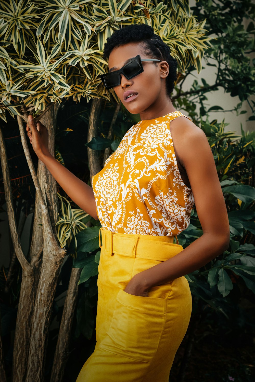 a woman wearing a yellow dress and sunglasses standing next to a tree