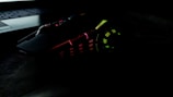 A gaming mouse with multicolored LED lights, including red, green, and yellow, has an angular, futuristic design. It sits next to a laptop keyboard in a dark setting.