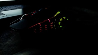 A gaming mouse with multicolored LED lights, including red, green, and yellow, has an angular, futuristic design. It sits next to a laptop keyboard in a dark setting.