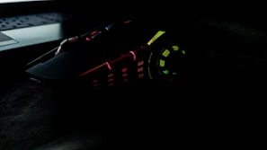 A gaming mouse with multicolored LED lights, including red, green, and yellow, has an angular, futuristic design. It sits next to a laptop keyboard in a dark setting.