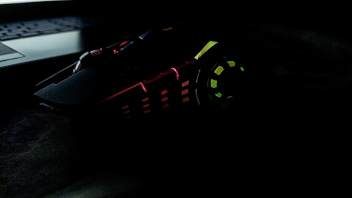 A gaming mouse with multicolored LED lights, including red, green, and yellow, has an angular, futuristic design. It sits next to a laptop keyboard in a dark setting.