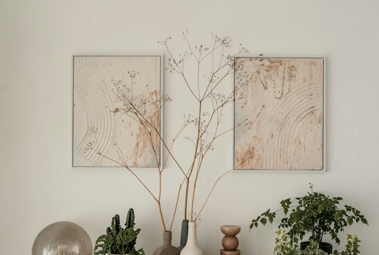A minimalistic arrangement featuring two abstract paintings with gentle circular patterns in earthy tones, accompanied by potted plants and vases. The central focus includes delicate branches in a vase, flanked by additional greenery, creating a serene and harmonious composition.