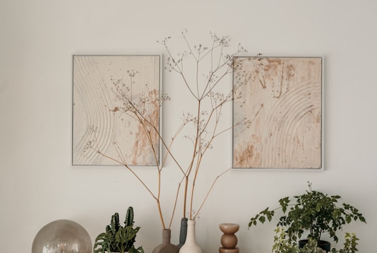 A minimalistic arrangement featuring two abstract paintings with gentle circular patterns in earthy tones, accompanied by potted plants and vases. The central focus includes delicate branches in a vase, flanked by additional greenery, creating a serene and harmonious composition.