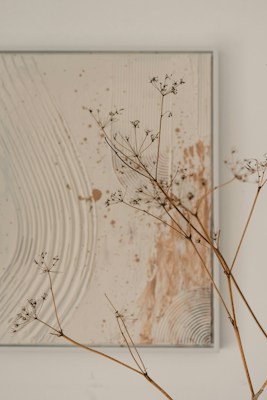 An abstract painting with swirling and splattered patterns in muted tones of beige and white dominates the background. In the foreground, delicate dried plant stems with small seed pods add an organic and natural element to the composition.