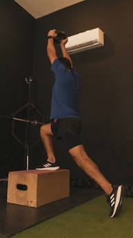 A person is performing a dynamic exercise. They are standing on one leg on a wooden box while holding a kettlebell overhead. The background is a dark gym with an air conditioning unit on the wall.