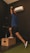 A person is performing a dynamic exercise. They are standing on one leg on a wooden box while holding a kettlebell overhead. The background is a dark gym with an air conditioning unit on the wall.