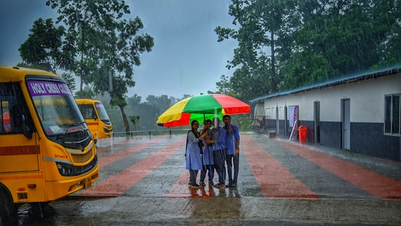 A group of people huddles under a brightly colored umbrella on a rainy day. Two yellow school buses are parked nearby, and the scene is set on a campus with a paved pathway and a building to the right. Tall trees surround the area, and the rain is visibly pouring down.