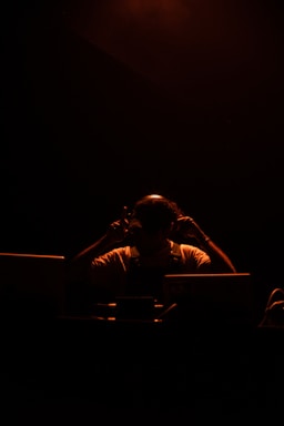 A person is silhouetted against a dark background, illuminated by a warm, soft light. The individual appears to be adjusting headphones, suggesting a focus on sound or music. The environment is dimly lit, creating a dramatic and intimate atmosphere.