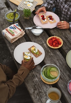 Various ingredients and snacks are arranged on a wooden table outdoors. There are plates with crispbreads topped with avocado, boiled eggs, cream, and strawberries. A bowl of sliced cucumbers, a jar of guacamole, and a small container of red berry slices are also present. Two people are interacting with the food, one holding a plate with a crispbread and egg.
