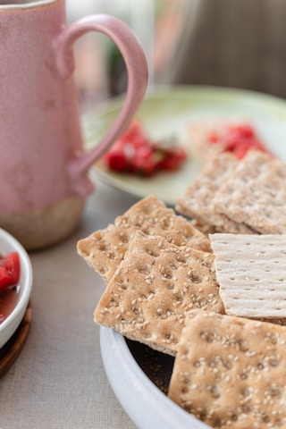 A close-up view of a plate with sesame seed-topped crackers in the foreground. Behind them, there is a pink ceramic mug and a plate with a serving of sliced strawberries, adding a splash of red color. The setting suggests a leisurely breakfast or snack on a table covered with a light beige tablecloth.