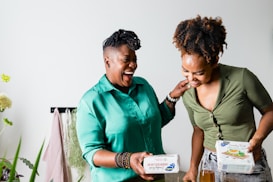 Two people standing side by side, engaged in a joyful moment. They are holding packages of crackers and appear to be sharing a conversation. The setting includes a few plants and a light-colored wall, creating a relaxed and casual atmosphere.