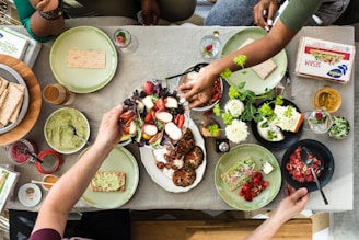 A table is filled with various food items, including plates of vegetables, slices of bread, and containers of hummus or dips. Several hands are seen reaching for the food amidst small bowls, glasses, and decorative plants.