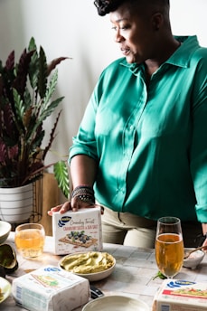 A person wearing a green shirt is standing near a table with various food items. There is a plant in the background, and the person is holding a box labeled 'Crunchy Twist Cranberry Seeds & Sea Salt'. Several drinks, a bowl of dip, and some avocados are placed on the table.