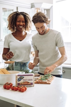 A man and a woman are in a bright kitchen, preparing food together. The woman is smiling and the man is concentrating on placing greens on a dish. The countertop is filled with ingredients including cherry tomatoes, a box of Wasa multi-grain crispbread, and some herbs in jars and containers.