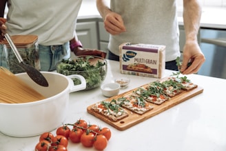 Two people are preparing a meal in a kitchen. There is a pot with dry spaghetti, a jar with herbs, and a bowl of greens on the counter. A package of multigrain crispbread is visible, along with a cutting board topped with crispbread, tomato sauce, cheese, and arugula. A bunch of tomatoes is placed in the foreground.
