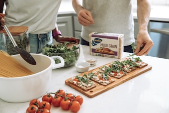 Two people are preparing a meal in a kitchen. There is a pot with dry spaghetti, a jar with herbs, and a bowl of greens on the counter. A package of multigrain crispbread is visible, along with a cutting board topped with crispbread, tomato sauce, cheese, and arugula. A bunch of tomatoes is placed in the foreground.